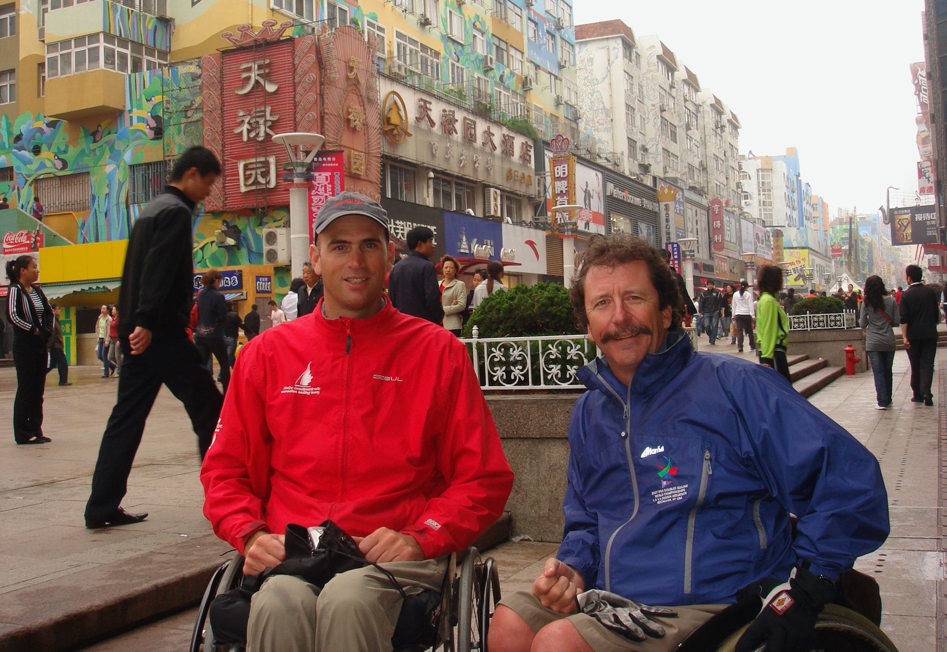 Paul Tingley and Danny McCoy in wheelchairs on a sidewalk in China with buildings in background