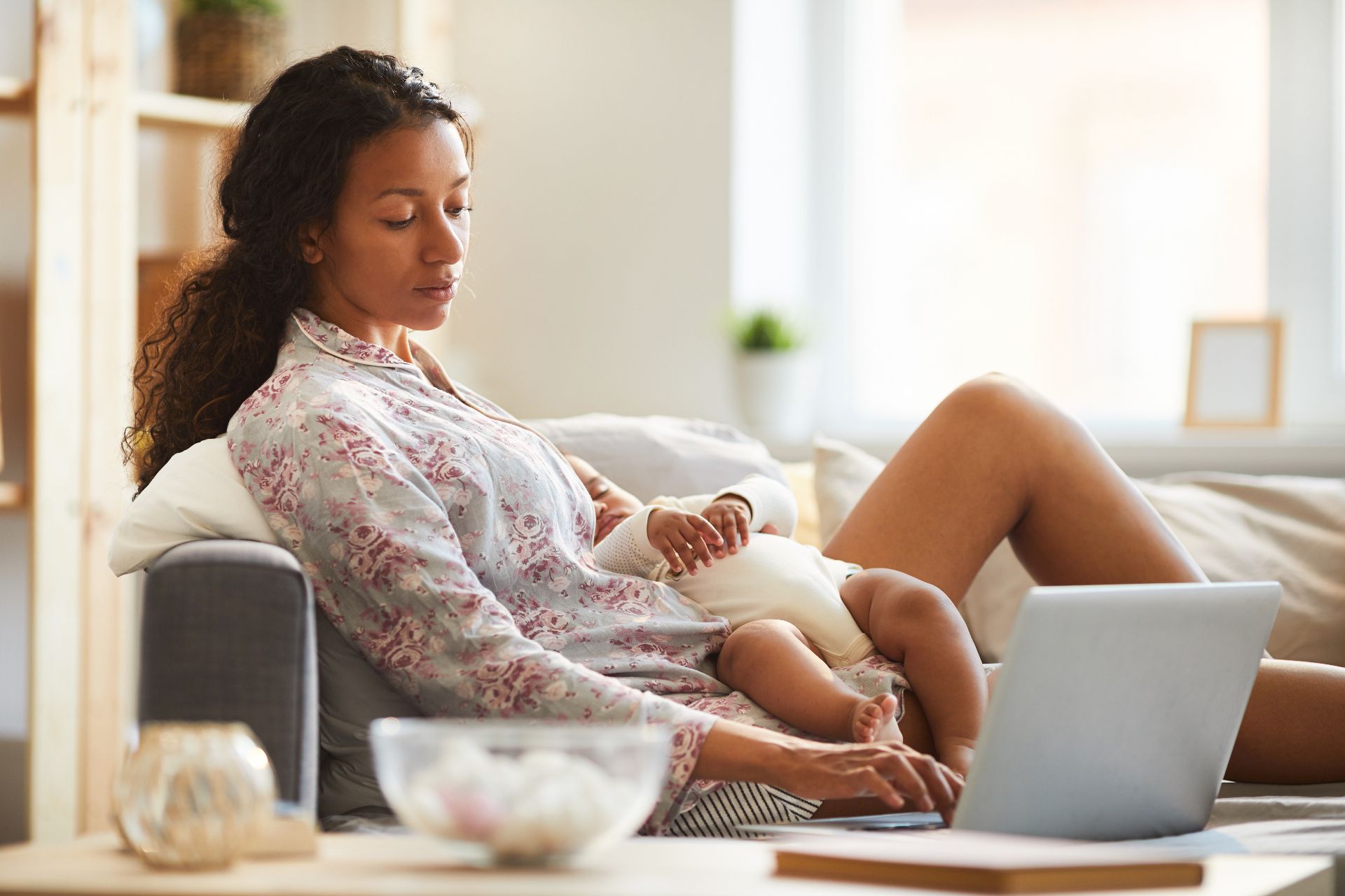 Black mother sitting on a couch with a sleeping baby while using a laptop at home, conveying the quiet emotional and physical demands of postpartum life.